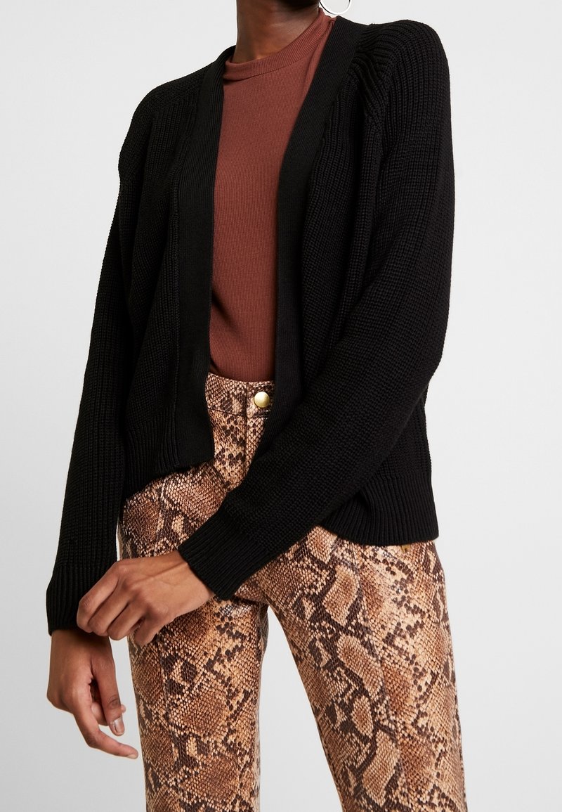 Personne portant un cardigan en tricot noir sur un haut marron, associé à un pantalon taille haute à imprimé serpent, vue du cou jusqu'aux hanches.