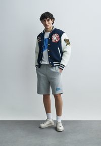 Ναυτικό μπλε και λευκό varsity μπουφάν με μπάλες, λευκό μπλουζάκι, γκρι σορτς και ανοιχτά αθλητικά παπούτσια. Οι κάλτσες είναι λευκές, το ύφασμα είναι βαμβακερό μείγμα.