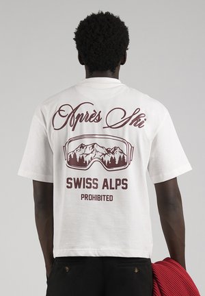 Un homme vu de dos porte un t-shirt blanc imprimé de lunettes de ski, paysage de montagne, et « Swiss Alps Prohibited », tenant un vêtement rouge.