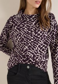 Lilla og creme mønstret langærmet skjorte med høj hals og tekstureret design, parret med sorte jeans med en metallisk knap.