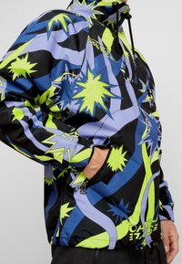 Personne portant une veste à capuche vibrante avec des motifs abstraits en éclat d'étoile et des rayures ondulées en néon vert, bleu et noir, la main dans la poche zippée.