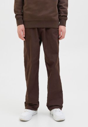 Personne portant un pantalon marron et un sweat-shirt marron assorti avec des baskets blanches, debout devant un fond blanc uni.