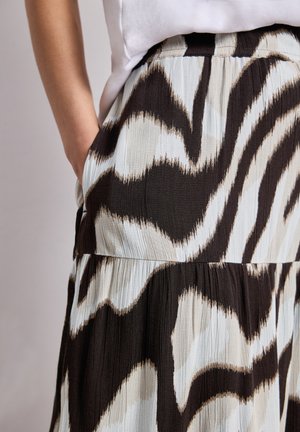 Kvinde iført plisseret nederdel med abstrakt brun, beige og hvidt mønster, hånd afslappet placeret i nederdelslomme, hvid top synlig.