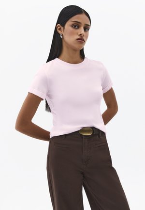 Donna con capelli lunghi e scuri che indossa una maglietta a maniche corte rosa chiaro, pantaloni marroni con fibbia in ottone e orecchini pendenti d'argento, in piedi con le mani dietro la schiena.