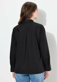 Camisa negra de manga larga con cuello, que presenta una espalda plisada, puños abotonados y una textura suave. Combinada con jeans de color azul claro.