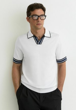 Muž s čiernymi okuliarmi, biela košeľa s krátkym rukávom, námornícky modrý pruhovaný golier a manžety na rukávoch, čierne nohavice, stojaci pri bielej stene.