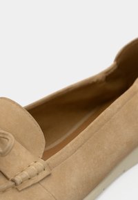 Mocassins en daim beige avec un bout rond, des détails cousus et un accent nœud sur le côté. Texture douce et design minimaliste.