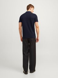 Mees seljaga pöördunud, kandes tumesinist lühikese varrukaga polo särki, musti viisakat pükste ja musti kingi, lihtsa heleda tausta ees.