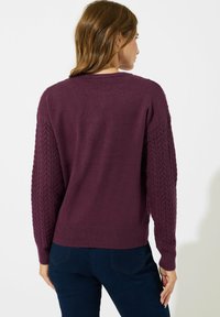 Maglione lavorato a maglia di un viola scuro con maniche a treccia, orlo a costine e colletto rotondo. Presenta un design fitted, leggermente cropped nella parte posteriore.