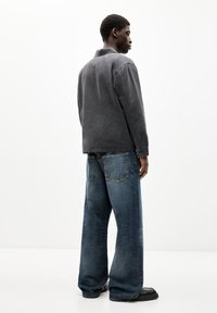 Übergroße graue Jacke mit lockerer Passform, getragen über blauen, weit geschnittenen Jeans. Schwarze, strukturierte Schuhe runden das Outfit ab.