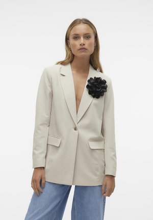 Blazer beige con bottone singolo, due tasche frontali e un accento floreale nero sul lato sinistro; tessuto liscio, design su misura.