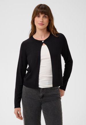 Femme portant un cardigan court noir avec un seul bouton en haut, par-dessus un haut blanc et un jean gris foncé, debout devant un fond uni.