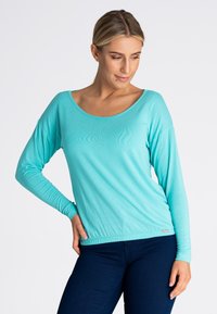 Turkusowa bluzka z długim rękawem wykonana z lekkiego materiału, z dekoltem w serek i marszczonym dołem, zestawiona z ciemnoniebieskimi dżinsami.