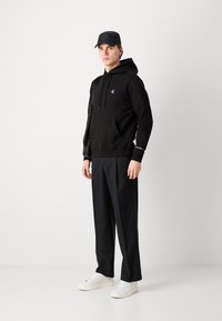 Haut à capuche noir avec une poche avant, logo blanc et accents aux poignets ; associé à un pantalon noir ample et des baskets blanches, le tout sur un fond neutre.