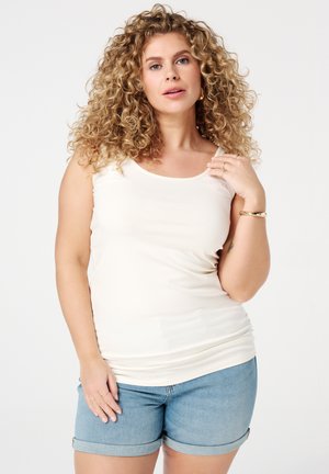 Femme aux cheveux blonds bouclés portant un haut blanc sans manches et un short en jean retroussé, debout devant un fond blanc uni.