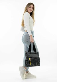 Black tote bag with textured fabric, dual black handles, two front zip pockets, and a yellow zipper pull. Model styled with light jeans and sneakers.