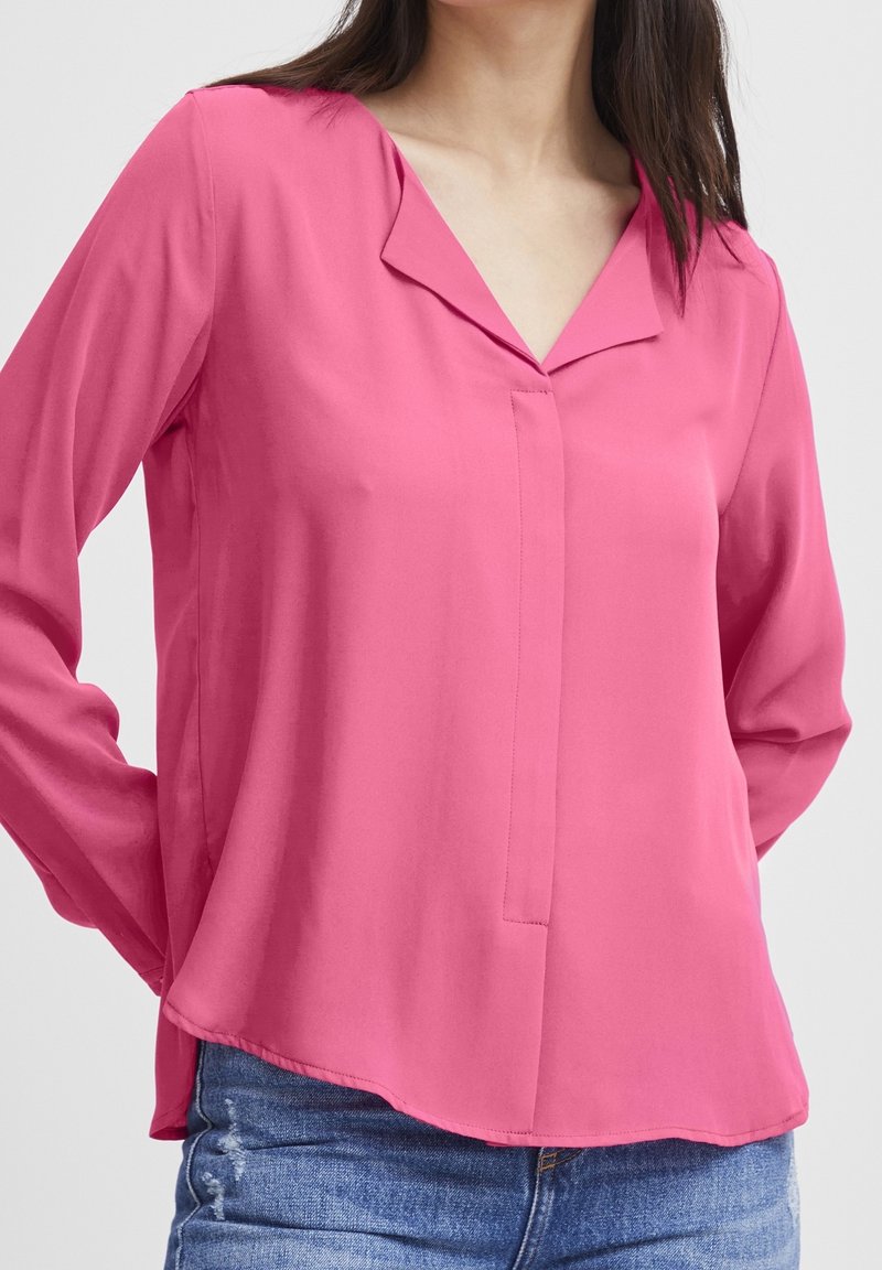 Femme portant un chemisier rose vif à manches longues avec un col en V et un jean bleu sur un fond clair uni.