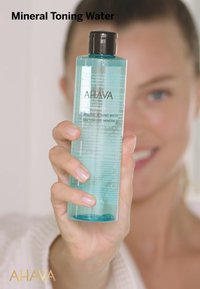 Przezroczysta plastikowa butelka AHAVA Mineral Toning Water, z turkusowym płynem, czarną zakrętką i teksturowaną etykietą z informacjami o produkcie oraz brandingiem.