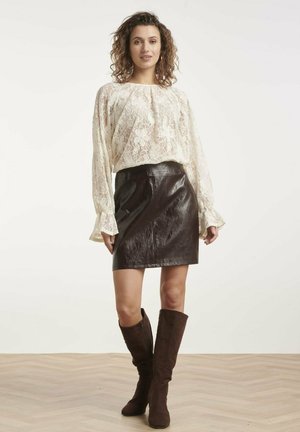 Vrouw die een witte kanten blouse, een zwarte leren rok en bruine kniehoge laarzen draagt, staand op een houten vloer tegen een witte achtergrond.