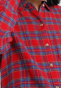 Röd rutigt skjorta med knäppning som har ett mönster av blå och vita linjer, rundad krage och pärlliknande knappar fram.
