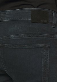 Vaqueros de mezclilla negra con una superficie texturizada. Presentan un parche negro sólido en la cintura con el nombre de la marca "DRYKORN". Se ve el bolsillo trasero.