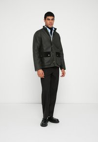 Oliwkowo-zielona kurtka użytkowa z czarnymi akcentami na rzepy i zatrzaskami, noszona na jasnoniebieskiej koszuli, zestawiona z ciemnymi spodniami i czarnymi butami.