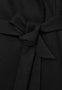 Must mustri vöö, mis on seotud sõlmesse villase jope külge, nähtava õmblusdetailiga.