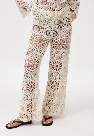 Crochet wide-leg pants in off-white, featuring hexagonal patterns with floral motifs and openwork detailing. Paired with black flip-flops.