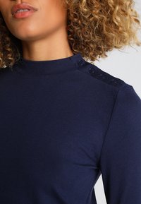 Maglietta a maniche lunghe a coste blu navy con colletto alto e dettagli di bottoni sulla spalla. Il tessuto è liscio ed elasticizzato, offrendo un look aderente.
