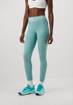 Woman wearing turquoise Nike Pro leggings, matching turquoise sneakers, white socks, and a white sports bra against a plain background.