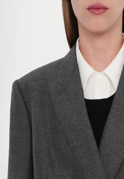 Blazer grigio in lana con rever a taglio, sovrapposto a una camicia bianca con bottoni e a un maglione nero a trama. Inquadratura ravvicinata che enfatizza i materiali.