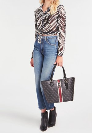 Kobieta ubrana w czarno-białą przezroczystą bluzkę w paski zebry, zawiązaną w talii, niebieskie dżinsy, czarne botki, trzyma czarną torebkę z wzorem i czerwonym paskiem.