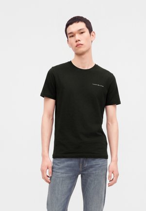 Млад мъж, облечен с черна тениска с къси ръкави Tommy Hilfiger и сини дънки, стои с неутрално изражение пред обикновен фон.