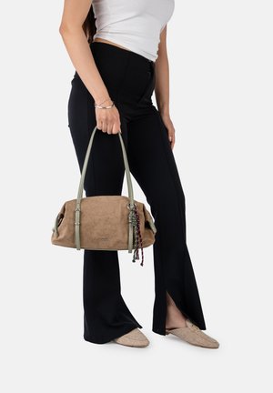 Frau in schwarzen Hosen und weißem Oberteil hält eine braune Wildleder-Handtasche mit grünen Trägern und geflochtenem Quaste, trägt beigefarbene Slipper.