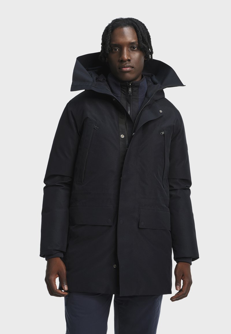 Man wearing a black winter parka with a high collar and multiple zippered and buttoned pockets, standing against a plain background.