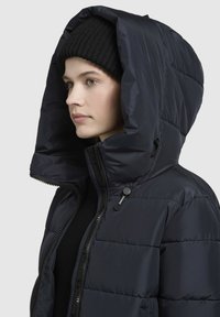 Tengeréskék steppelt dzseki nagy, estruturált kapucnival, elől cipzárral és varrási részletekkel. Fekete kötött sapka van alatta.