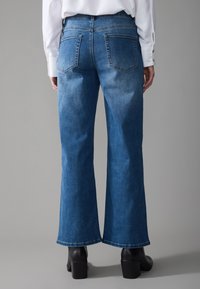 Wide-leg denim jeans in medium blue wash, featuring two back pockets, subtle fading, and a raw hem. Styled with black block-heeled boots.