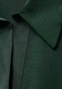 Blusa verde oscura con una textura suave, que presenta un cuello clásico y costuras precisas a lo largo de la tapeta. La tela parece ligera.