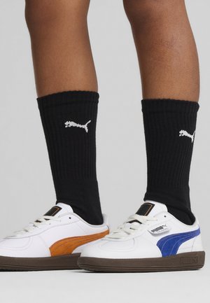 Zapatillas Puma de cuero blanco, con rayas naranjas y azules, suela de goma texturizada en marrón, combinadas con calcetines negros de canalé que llevan un logo blanco.