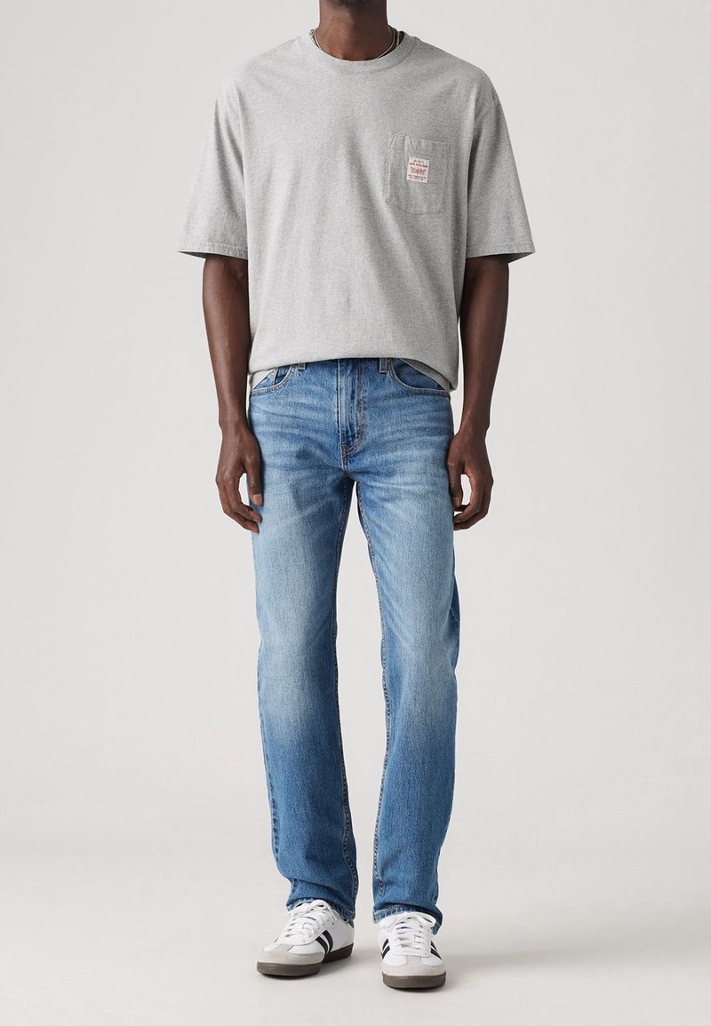 Homme portant un t-shirt gris clair à manches courtes avec une poche et un jean bleu clair, debout devant un fond uni.