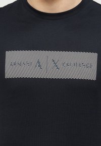 Μαύρο μπλουζάκι με στρογγυλή λαιμόκοψη και λευκό διαγώνιο ριγέ ορθογώνιο με το κείμενο «ARMANI A | X EXCHANGE» στο στήθος.