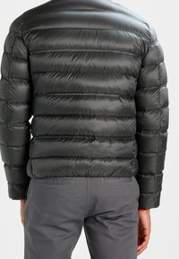 Zwarte pufferjack met horizontaal gestikt ontwerp, glanzende textuur en een gladde kraag. Gecombineerd met grijze broek, gezien vanuit de achterkant.