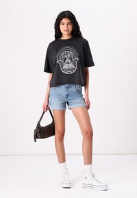 Juodas trumpas T-shirt su balta grafika; derinamas su šviesiai mėlynomis džinsinėmis šortais. Modelis laiko mažą rudą krepšį, dėvi baltus sportbačius.