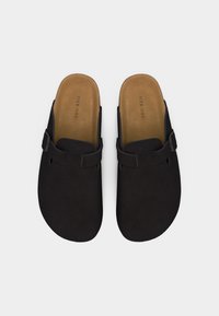 Schwarze Slip-On-Clogs mit braunen Innensohlen und verstellbaren Riemen mit Schnalle, von oben auf weißem Hintergrund gezeigt.