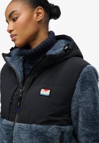 Czarna i niebieska kurtka puchowa z kapturem o teksturze polarowej, z zapięciem na zamek błyskawiczny i naszywką z logo. Posiada żebrowany kołnierz oraz miękką podszewkę.