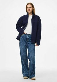 Marinblå fleecejacka med hög krage och dragkedja framtill, kombinerad med ljusblå vida jeans och beiga skor.