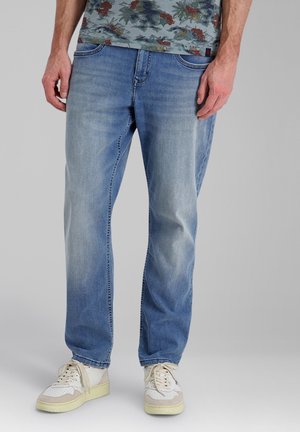 Mann trägt hellblaue Jeans mit lockerem Schnitt, weiße Turnschuhe und ein Hemd mit Blumenmuster, steht vor einem einfarbigen Hintergrund.
