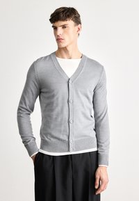 Sivi kardigan z V-izrezom, rebrastimi manžetami in robom, z šestimi gumbi spredaj, oblečen čez belo majico s kratkimi rokavi.