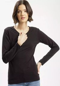 Czarna bluzka z długim rękawem i dekoltem w szpic, wykonana z gładkiego materiału, dopasowany krój i minimalne detale. Model pozujący z jedną ręką na piersi.
