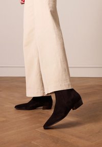 Bottines en suède noir avec un bout pointu et un petit talon carré, assorties à un pantalon large crémeux. Un parquet en bois en arrière-plan.